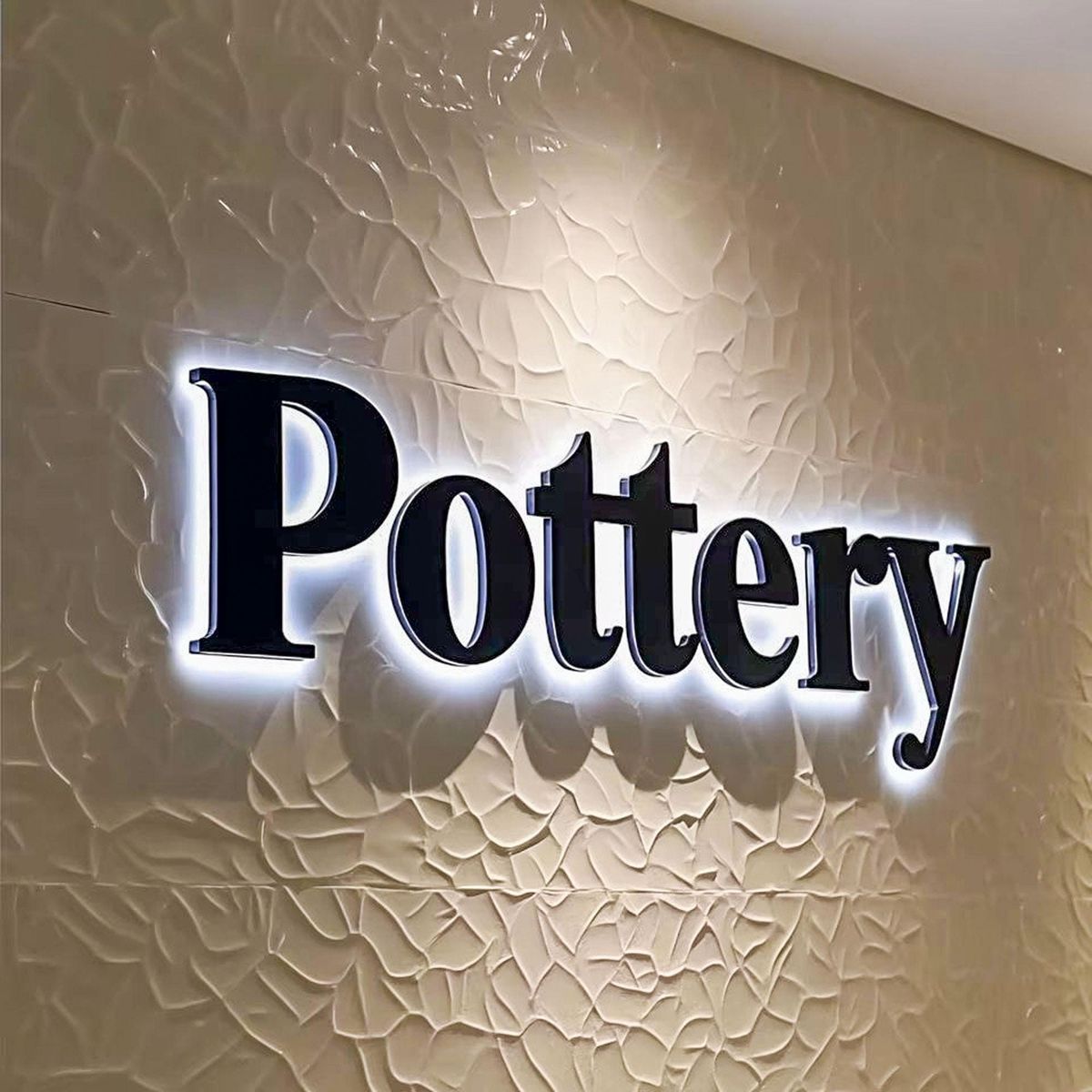 Custom Pottery acrylic letter sign with white halo LED backlighting mounted on a textured decorative wall.
