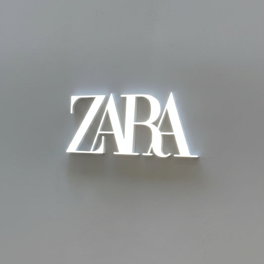 Custom Zara-style LED acrylic logo sign with bright white illumination mounted on a modern gray wall.